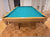 A wooden pool table with a bright teal green felt playing surface, photographed in a home interior with a wood parquet floor and white doors in the background.