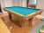 A wooden pool table with turquoise felt is shown in a home interior with wood floors and a mirror reflecting the table.
