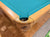 Close-up of a Duttlin pool table corner with a turquoise felt surface and a light natural wood frame, shown on a wood parquet floor.
