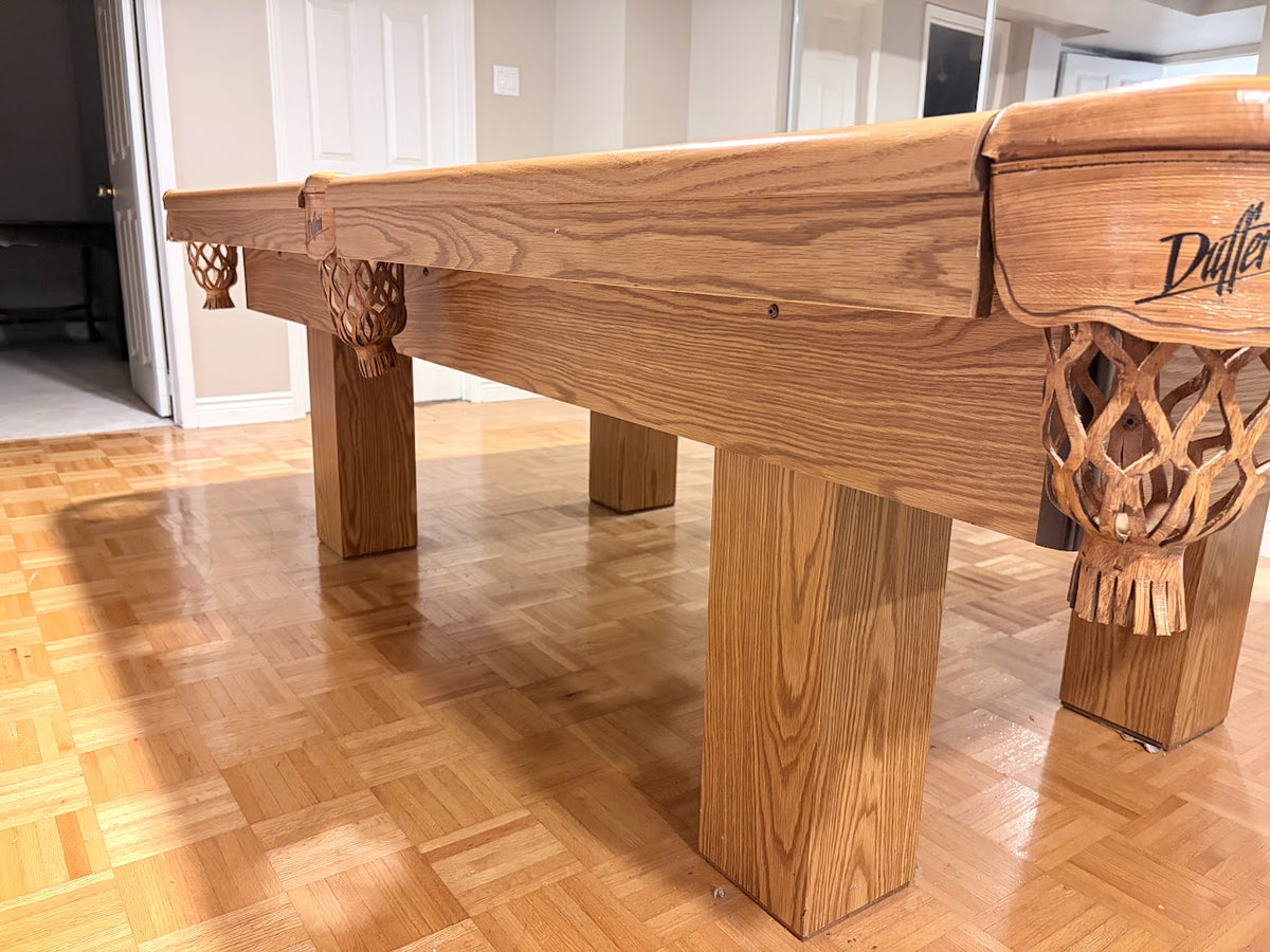 A close-up of a wooden pool table with a natural oak finish, featuring carved wooden gutters and legs, set on a polished wood parquet floor in an indoor room.
