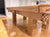 A close-up of a wooden pool table with a natural oak finish, featuring carved wooden gutters and legs, set on a polished wood parquet floor in an indoor room.