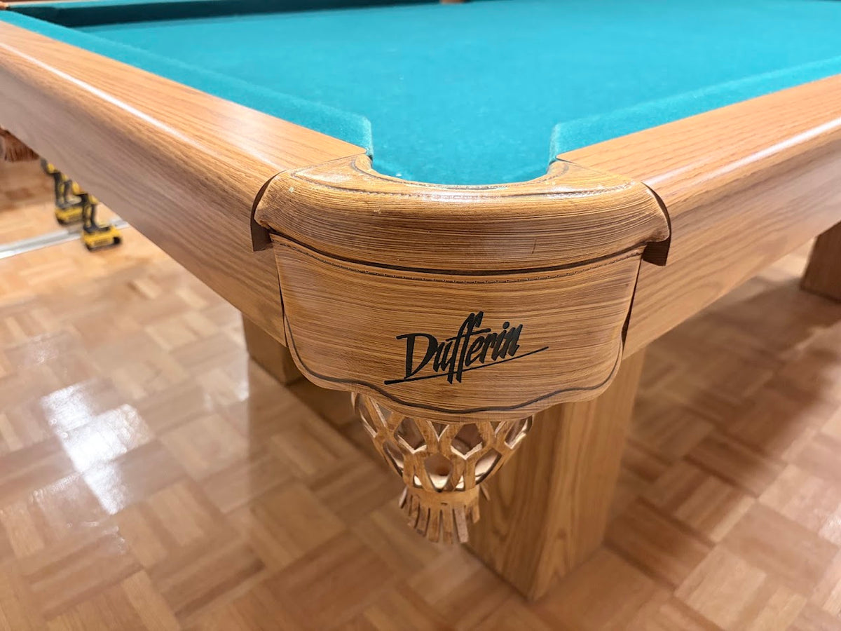 Close-up of a wooden Dufferin pool table corner with turquoise felt and a carved wooden cue pocket, shown on a wood parquet floor.