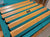 Six long wooden molding or trim pieces with a glossy light oak finish are arranged diagonally on a teal surface. A small metal plaque is attached to one piece, and a small tool is visible in the upper left corner.