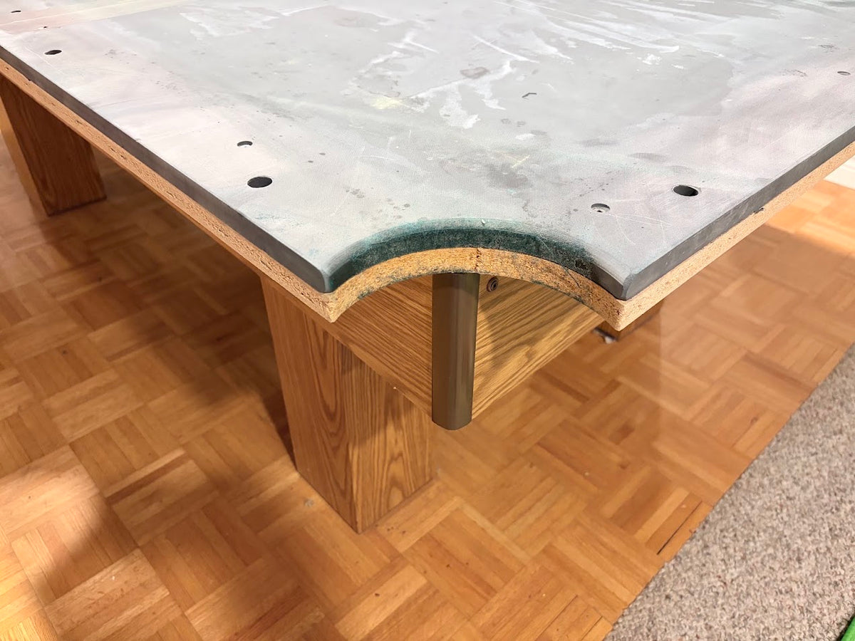 A close-up of a wooden table corner with a curved edge, featuring a light gray stone or concrete top, a medium brown wood base, and a dark metal support leg, set on a parquet wood floor.