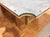 A close-up of a wooden table corner with a curved edge, featuring a light gray stone or concrete top, a medium brown wood base, and a dark metal support leg, set on a parquet wood floor.