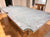 A large rectangular table with a thick, curved concrete top and light wood legs, photographed in a home interior with wood floors and other furniture visible in the background.