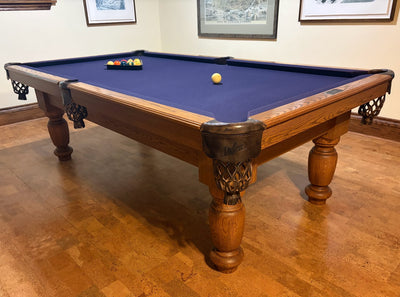 A wooden pool table with a blue felt playing surface, shown in a home game room with a wood floor and framed wall art.