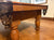 A close-up of a wooden pool table showing the side pocket, cue stick rest, ornate metal netting, and turned legs on a polished stone floor.