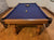 A wooden pool table with a dark blue felt surface, shown in a home setting with wood floors and a stone wall in the background.