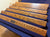 Six wooden stair rails with a natural oak grain pattern, arranged diagonally on a blue velvet-covered surface. A small metal brand plaque is attached to one of the rails.