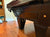 A close-up of a dark brown wooden pool table leg and side panel, showing carved shell details and brass hardware, set in a room with hardwood floors.