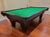 A dark wood pool table with a green felt playing surface, photographed in a room with a polished wood floor and beige walls.