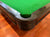 Close-up of a green felt pool table corner with a dark wood or laminate rail and a black pocket cup, set on a polished wooden floor.
