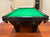 A black pool table with a green felt playing surface, photographed in a room with wood floors and beige walls.