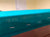 Close-up of a teal fabric-covered surface with the text 'IWAN SIMONIS 760' printed in light beige, along with small white dash marks, photographed indoors.