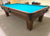 A wooden pool table with a bright turquoise blue felt surface, photographed in a carpeted indoor room with furniture visible in the background.
