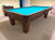 A wooden pool table with bright turquoise felt is shown in a home or club room setting with beige carpet and light yellow walls.