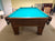 A wooden pool table with a bright turquoise felt surface, photographed in a room with carpet, a lamp, wall art, and storage boxes in the background.