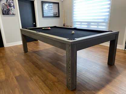 An 8' Majestic Mensa Pool Table with a black felt surface and dark brown wooden edges, placed on a hardwood floor in a room with a window and a picture on the wall.