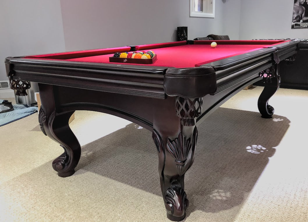 A  mahogany wood pool table with red cloth top, featuring carved leg designs and leather-fringed pockets and set of balls on the surface.