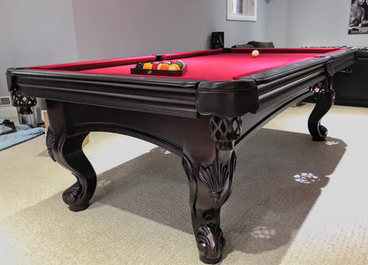 A  mahogany wood pool table with red cloth top, featuring carved leg designs and leather-fringed pockets and set of balls on the surface.