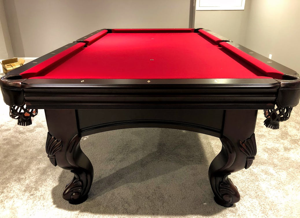 A  mahogany wood pool table with red cloth top, featuring carved leg designs and leather-fringed pockets.