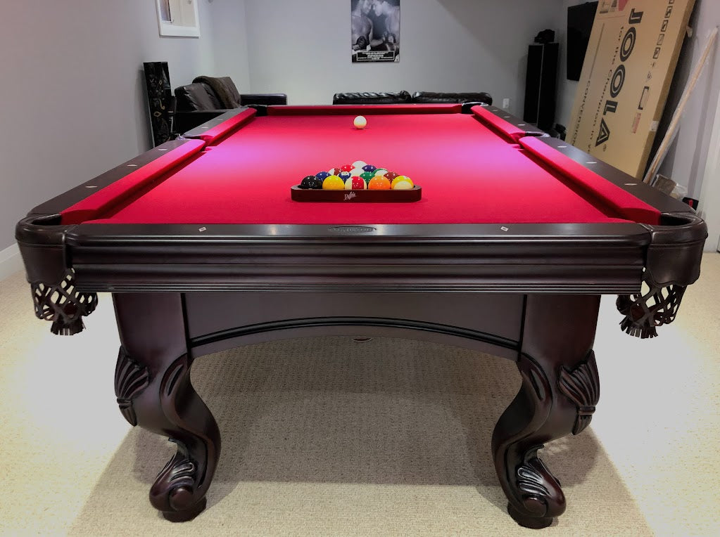 A  mahogany wood pool table with red cloth top, featuring carved leg designs and leather-fringed pockets and set of balls on the surface.