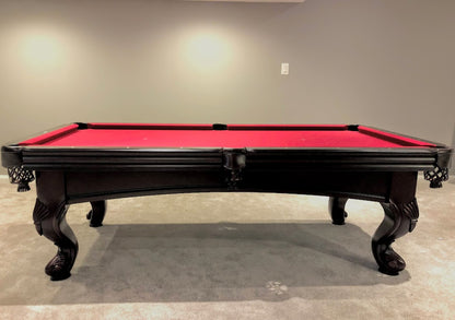 A  mahogany wood pool table with red cloth top, featuring carved leg designs and leather-fringed pockets .