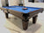 A 9' Majestic Ascot Walnut Pool Table with a blue felt surface, brown wooden frame and legs, and ball and cue placed on the table.
