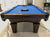 A 9' Majestic Ascot Walnut Pool Table with a blue felt , walnut wooden frame and legs, and ball and cue placed on the table.