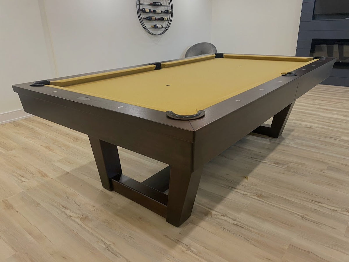 A sleek brown pool table with a yellow-finished playing surface, situated in a room with wooden flooring and a wall-mounted award or certificate.
