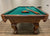 Billiard table in brown walnut finishing with green felt and balls and cues on the top and logo.
